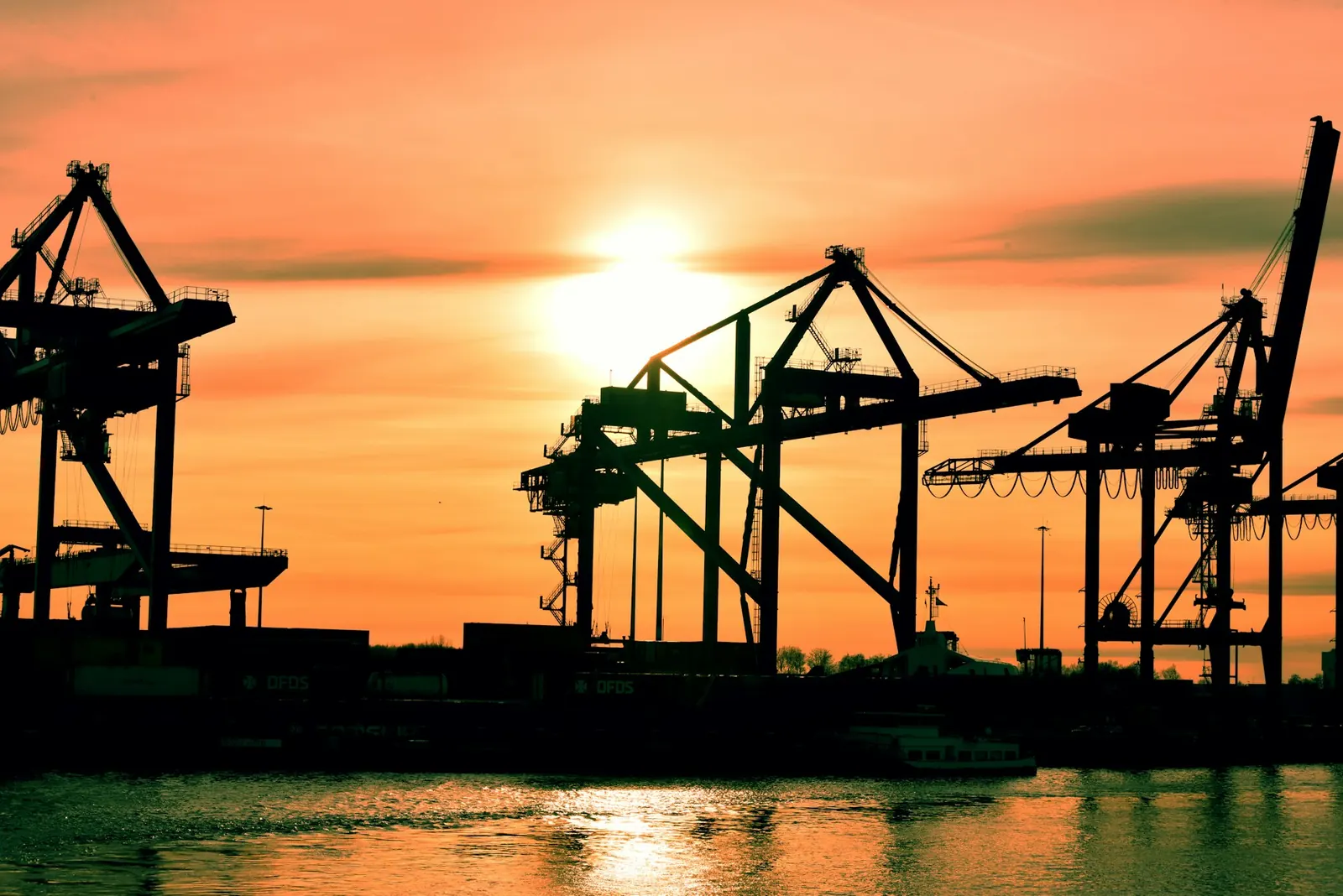 Silhouetted harbour cranes against an orange sunset over the water.